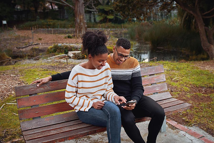 Client Center - Happy Young Couple Use a Phone on a Park Bench